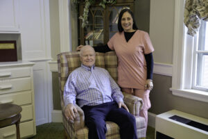 Caregiver in pink scrubs standing beside an elderly man seated in a plaid armchair in a cozy living room.