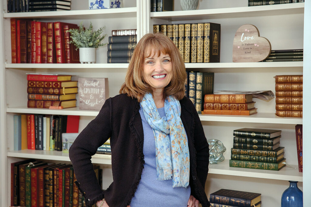 Elizabeth_Russell_TracyJenkins-4 - Saint Elizabeth Community Smiling woman in a blue sweater and light scarf stands in front of white bookshelves filled with colorful books.