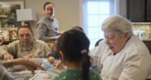 Seniors and a caregiver sit around a table doing a craft activity in a sunny dining area.