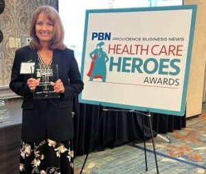 Woman in a black blazer and floral dress holds a glass award beside a sign for Health Care Heroes Awards.