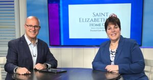 Two news hosts sit at a desk in a studio, with a screen behind them displaying the Saint Elizabeth Home logo.