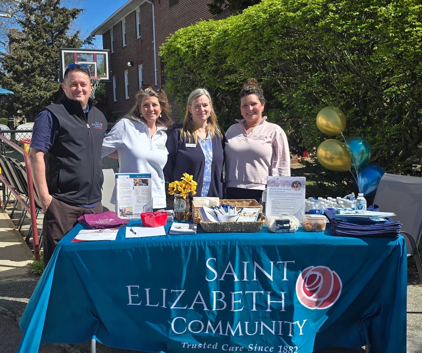 Four community volunteers stand behind a table with a Saint Elizabeth Community banner at an outdoor info booth. Balloons and brochures are on the table.