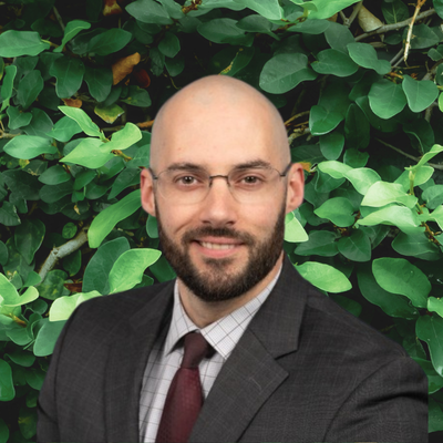 Head-and-shoulders portrait of a bald man with a beard and glasses in a dark suit and burgundy tie, posed in front of green foliage.