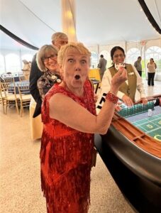 Older woman in a red sequined dress holds up a chip at a craps table, surprised expression, with other guests in the background under a tent.