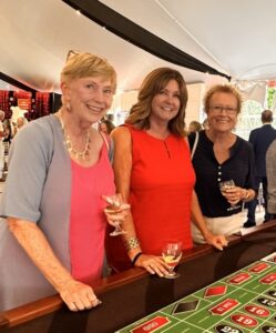 Three women stand at a casino gaming table, each holding a wine glass and smiling at the camera.