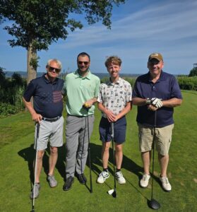 Four men posing with golf clubs on a sunny golf course, with trees and water in the background.