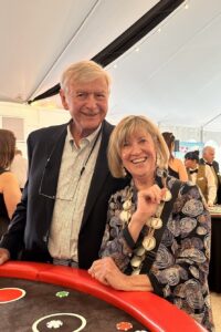 Smiling elderly couple posing at a casino table with poker chips on red felt under string lights.