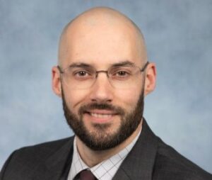 Professional headshot of a bald man with glasses and a beard in a dark suit, against a blue backdrop.