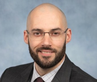 Professional headshot of a bald man with glasses and a beard in a dark suit, against a blue backdrop.