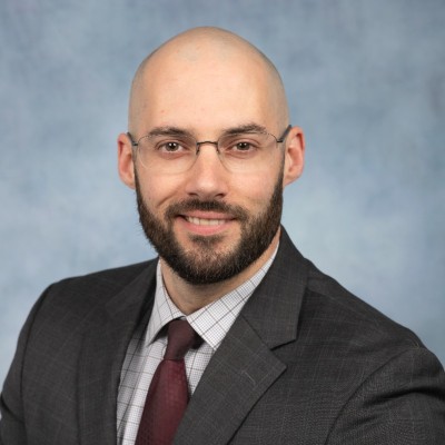 Professional headshot of a bald man with a beard and glasses, in a dark suit and burgundy tie, against a blue backdrop.