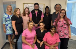 CNA Training Program June 2026 Graduates - Saint Elizabeth Community Group of diverse medical staff posing together in a clinic hallway, some wearing pink scrubs and others black scrubs, with a framed certificate on the wall behind them.