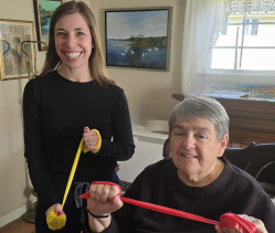 Smiling woman and older man sit indoors, holding colorful exercise sticks with soft balls at the ends, doing light arm stretches together.