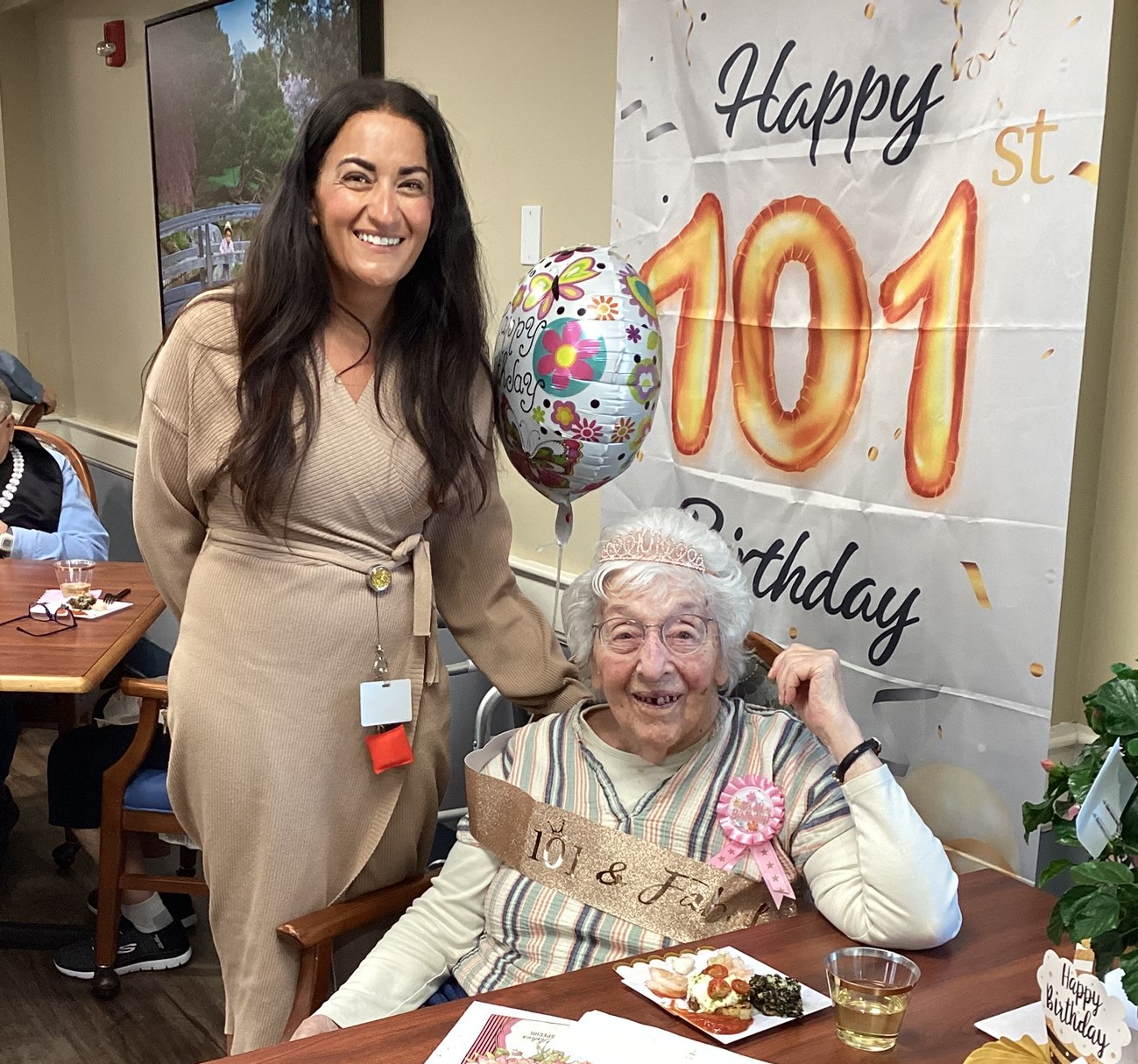 Young woman in a beige wrap dress stands beside an elderly lady with a tiara at a 101st birthday party.