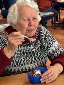 Older woman with short white hair eating dessert from a blue bowl with a spoon in a café-like setting