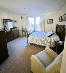 GVG-Room-scaled interior - Saint Elizabeth Community Bedroom with a dark wooden bed on the right, white-and-black patterned comforter, a dresser with a mirror and framed photos on the left, and a window with light curtains in the back.