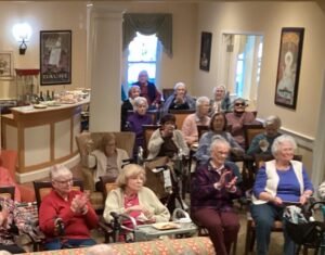 Group of elderly residents seated in a lounge, some in wheelchairs, clapping and enjoying a social event with a buffet visible in the background.