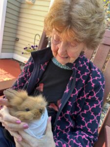 IMG_4660 - Saint Elizabeth Community An elderly woman wearing a patterned navy jacket sits on a porch while holding a fluffy piglet wrapped in a light cloth.