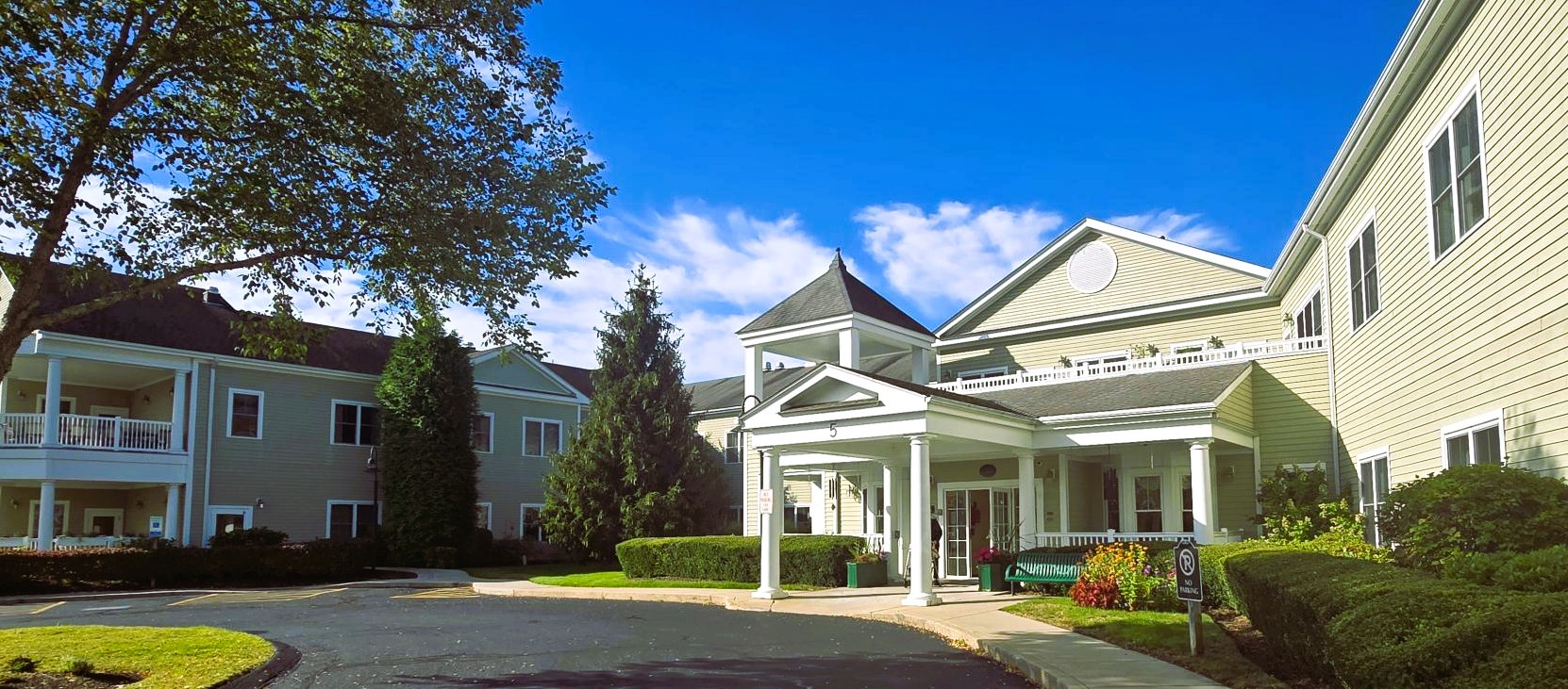 Exterior view of a beige multi-wing building with a white-columned covered entrance and well-manicured shrubbery under a bright blue sky.