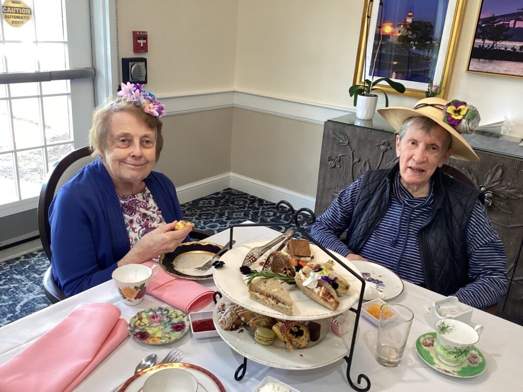 Two elderly women wearing flower hats sit at a table with a two‑tier dessert and sandwich stand, smiling at tea service.