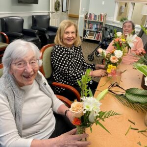 Three smiling elderly women sit at a table arranging fresh flowers in vases, with scissors and greenery on the table in a communal room.
