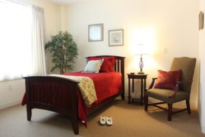 Cozy bedroom with a wooden bed, red quilt, and beige walls; lamp on the nightstand, armchair, and slippers on the carpet.