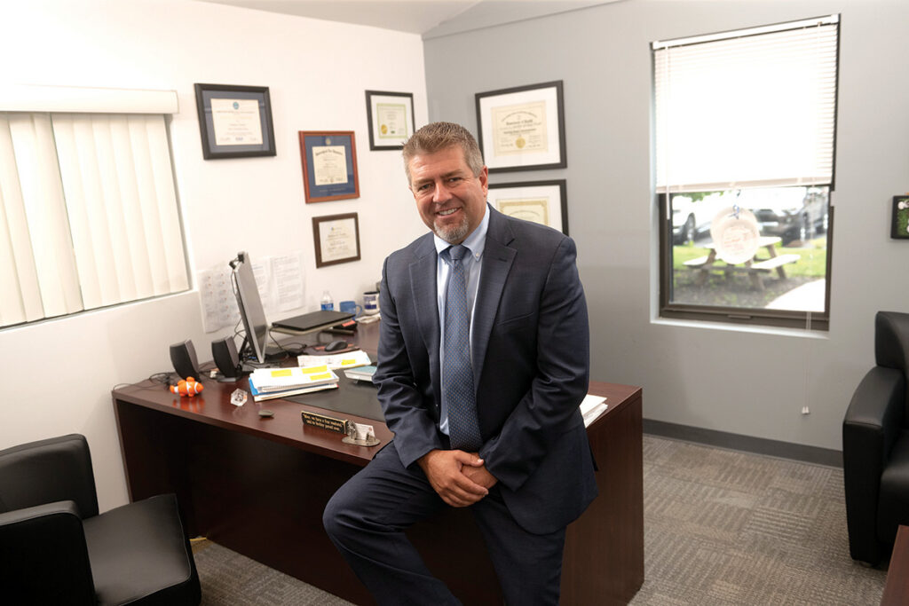 Smiling man in a navy suit and blue tie sits on the edge of a wooden desk in a tidy office with framed certificates on the wall.