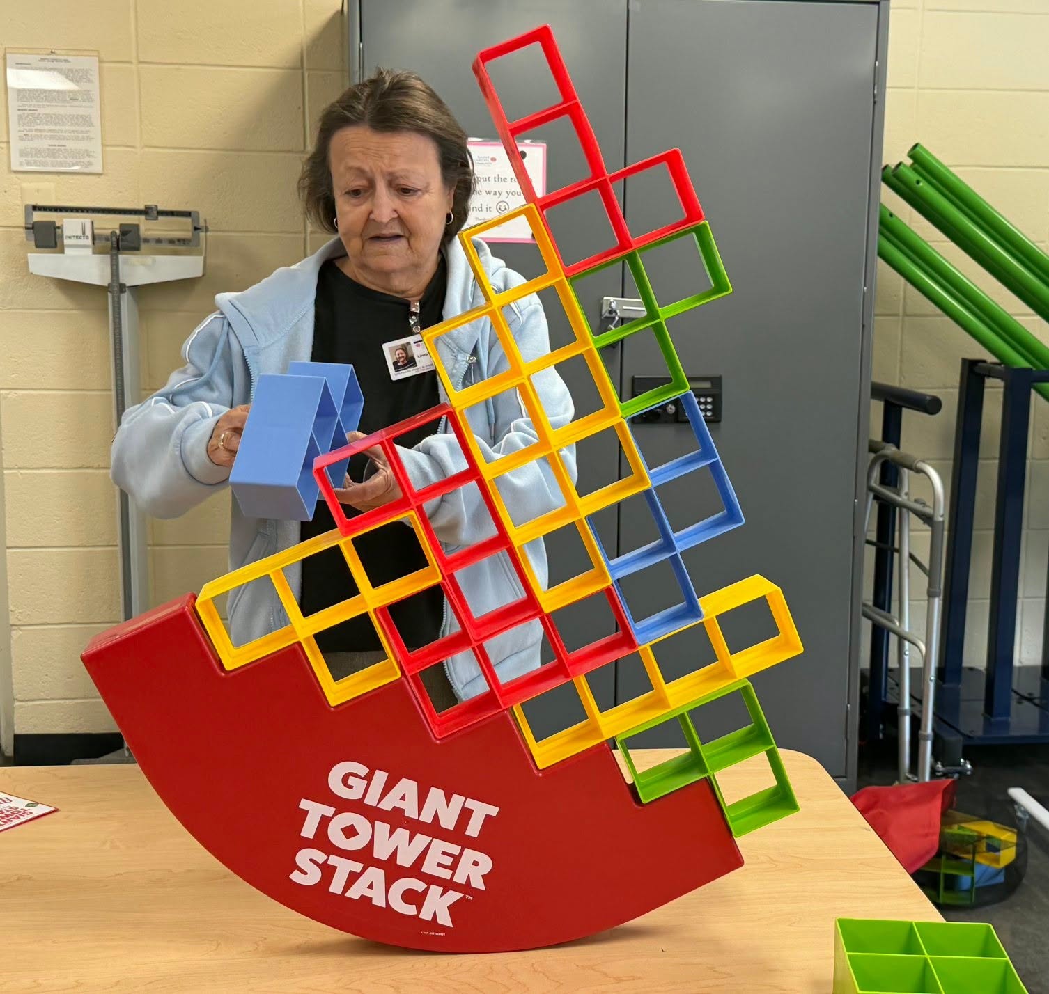 Woman assembling a large colorful plastic tower stack game, arranging red, yellow, green, and blue square frames while looking focused in a classroom setting