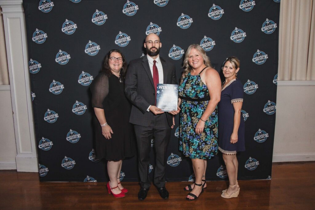 Group of four adults posing together at an awards event; man in suit holds a 'Winner' booklet, backdrop displays Readers' Choice logos.