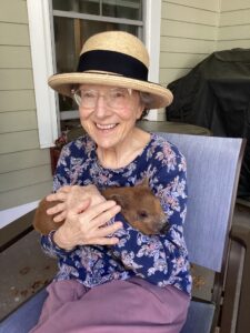 IMG_0647 - Saint Elizabeth Community Smiling elderly woman wearing a straw hat and glasses, cradling a small brown piglet on her lap on a porch.