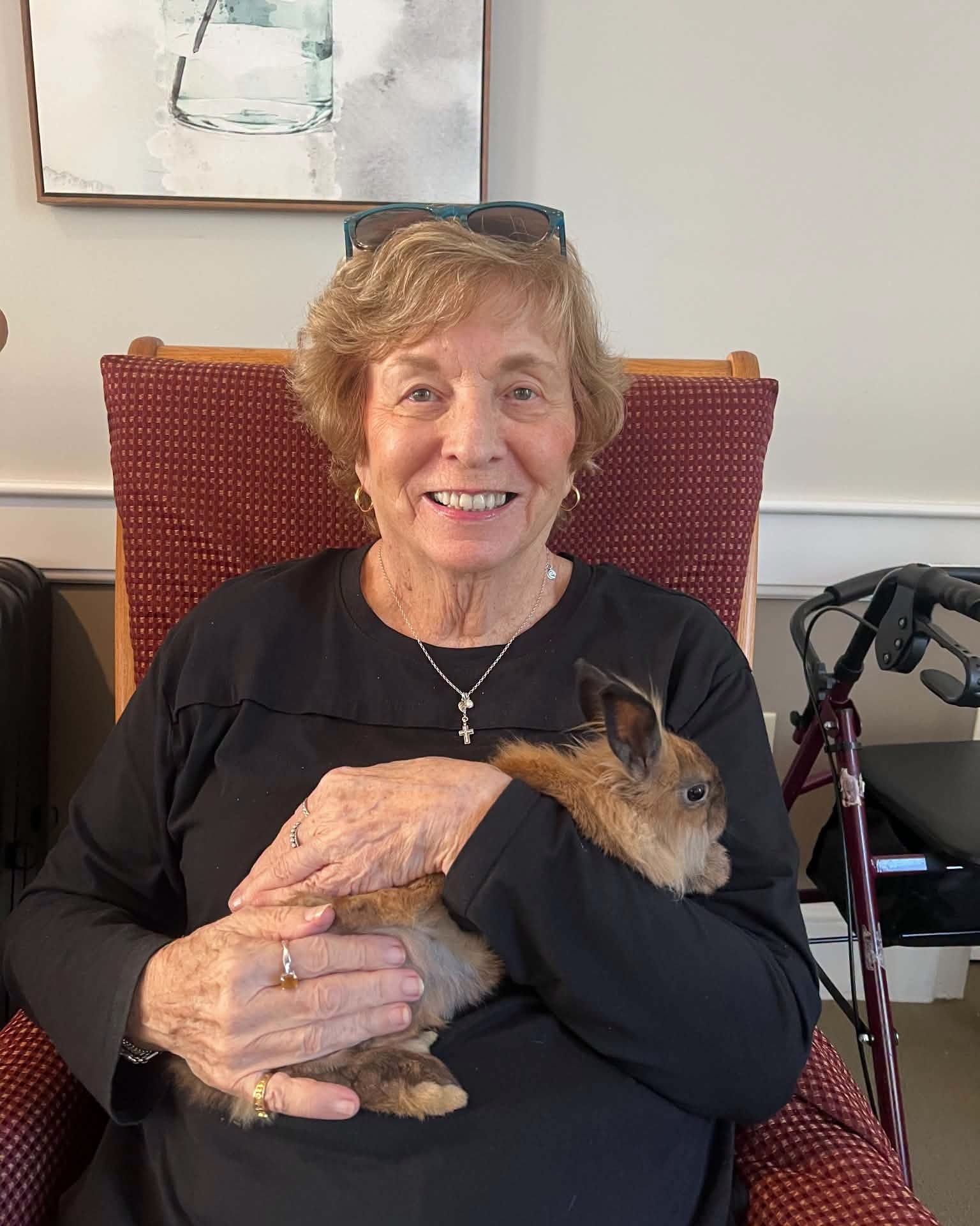 Smiling elderly woman with short blond hair, glasses on her head, holding a brown rabbit.