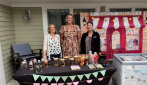 Three women stand behind a table filled with jars of jams and treats at a Sweet Shoppe booth.