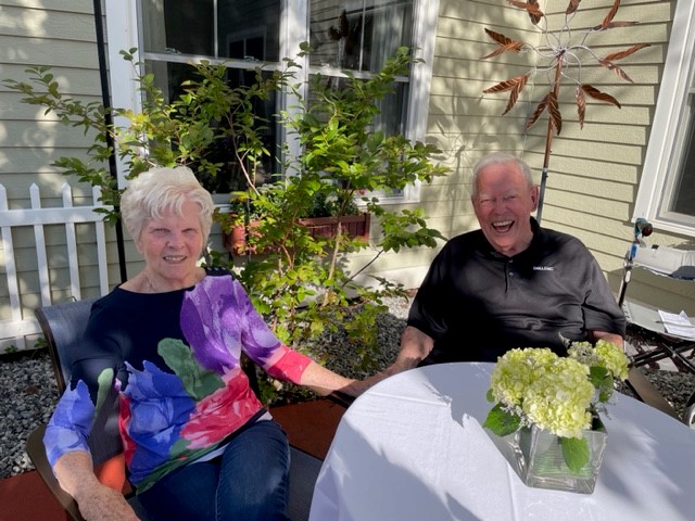 Older couple sitting at a round table outside, smiling, with greenery and a beige house in the background; a small flower arrangement sits on the table in a glass vase.