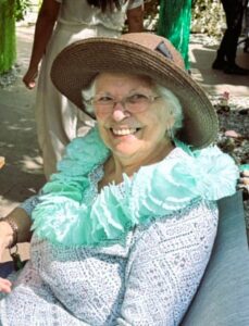 Smiling elderly woman wearing a wide-brim straw hat and mint-green ruffled scarf, seated outdoors.