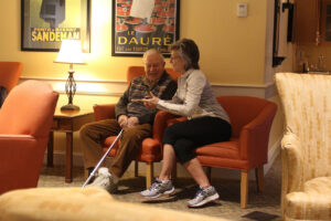Elderly man with a cane sits in an orange chair while a woman chats with him in a cozy living-room lounge with posters on the wall and a lamp nearby.